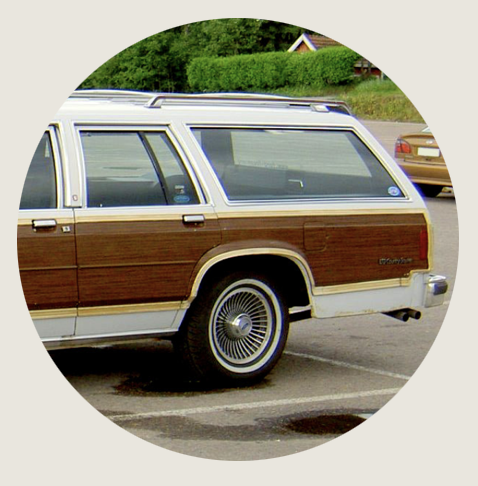 A classic brown-and-white wood-paneled station wagon in a parking lot. The car’s long body, chrome accents, and spoked hubcaps evoke mid-century Americana nostalgia. Trees and another car sit in the background.
