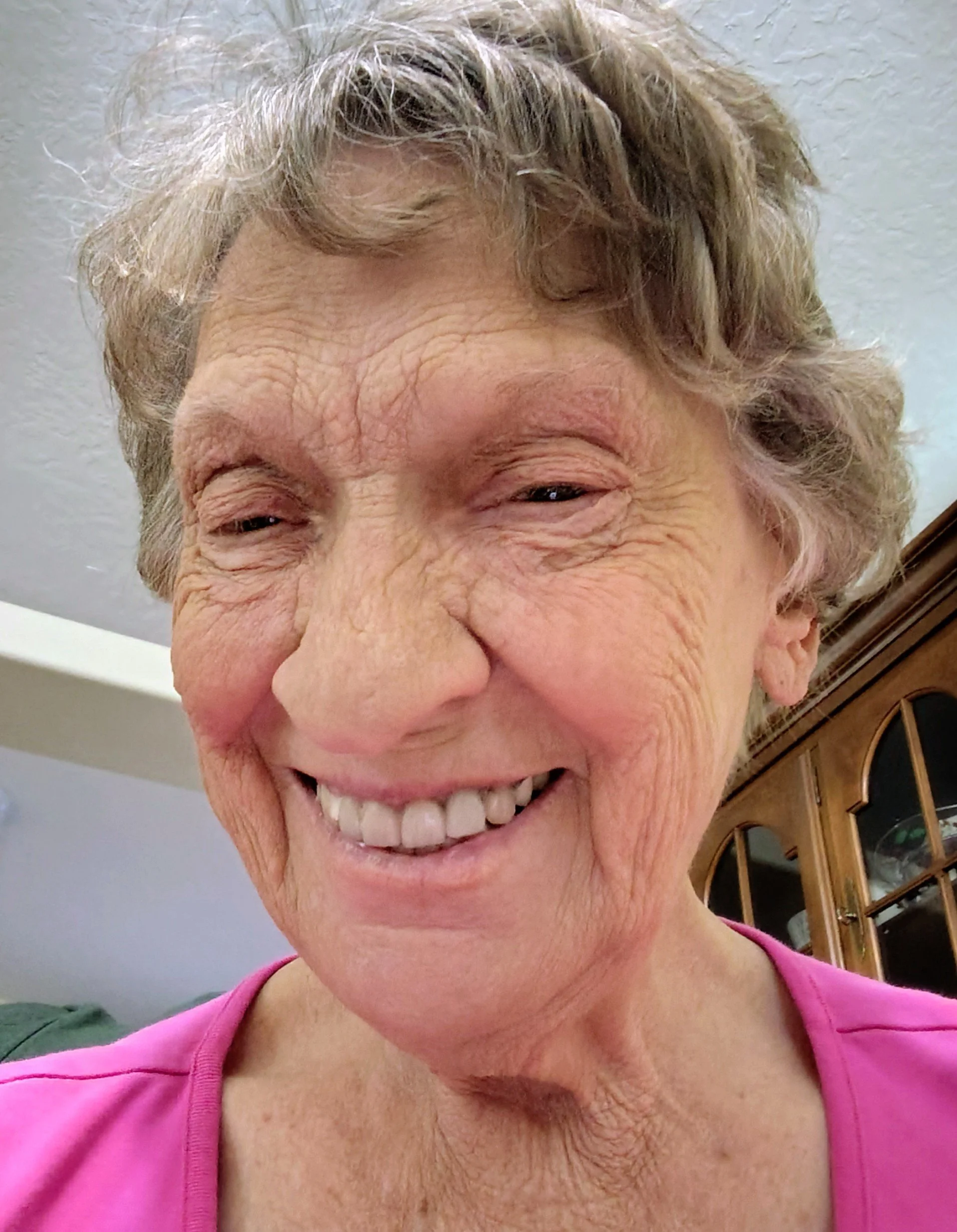 A close-up portrait of an elderly woman with short, wavy gray hair, smiling warmly while wearing a bright pink shirt