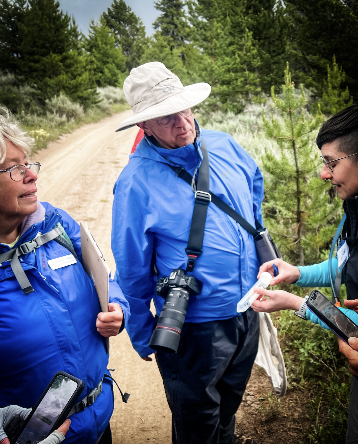 Kurt Wambier in the foreground in the middle of the photo. He wears a tan sun hat, a blue raincoat, and has a camera draped over his left shoulder. A person flanks him on his right and left. The person on his left is holding a specimen tube.