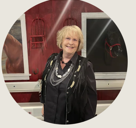 Cheyenne- A smiling woman with blond hair stands in front of a red wall decorated with horse-themed paintings. She is dressed in black with fringe and layered necklaces.