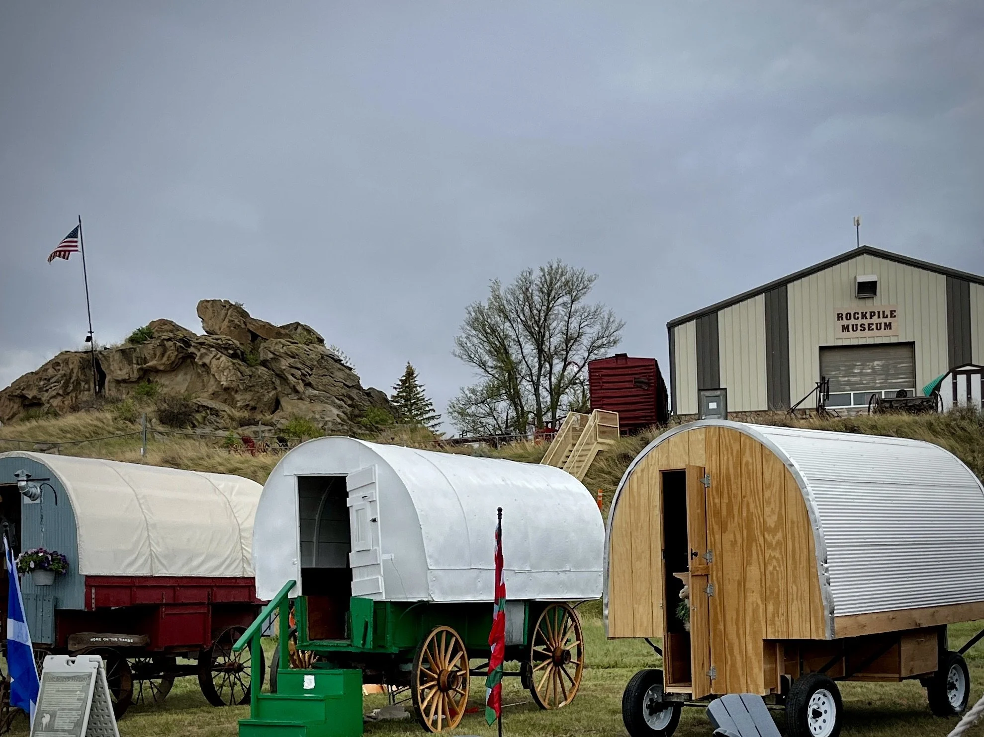 Three covered sheep wagons are parked in front of the Rockville Museum, behind them to the right. Behind them to the left is a pile of rocks with an American Flag anchored in.