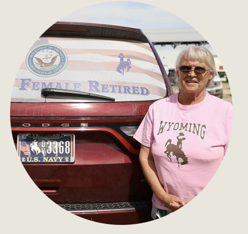 A woman in a pink “Wyoming” T-shirt stands beside a red Dodge SUV with a Navy veteran license plate. The back window displays a United States flag decal with “Female Retired” and a Wyoming bucking horse and Navy logo.