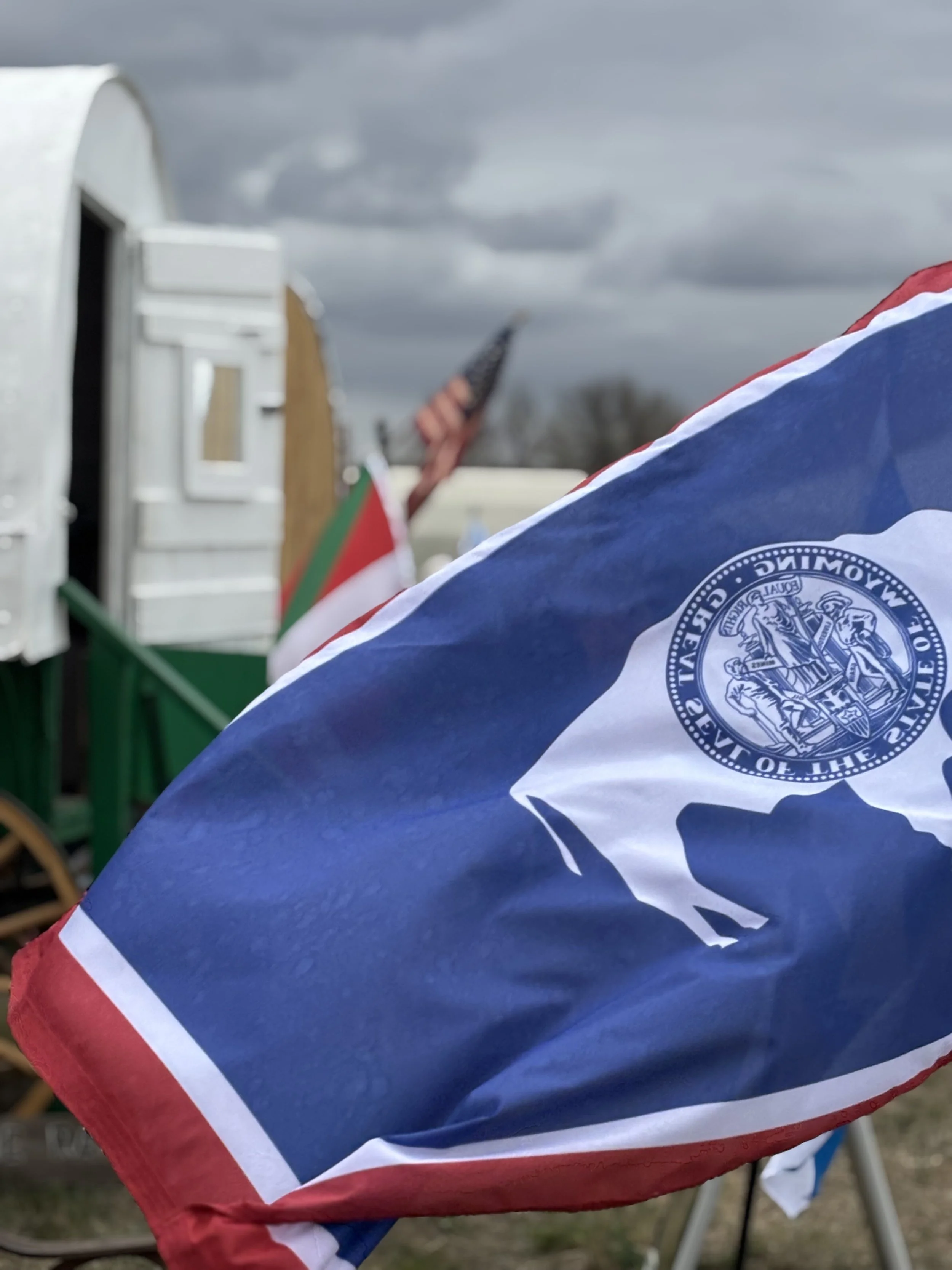 A close up of the Wyoming state flag in the foreground, with two out of focus sheep wagons and an American flag in the background.