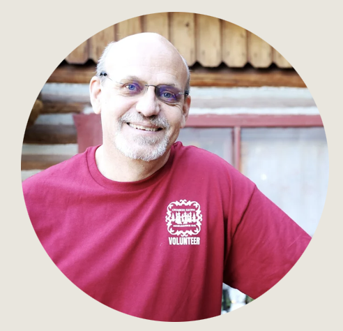 Casper- A man wearing glasses and a red “Volunteer” T-shirt smiles warmly in front of a rustic log building. 