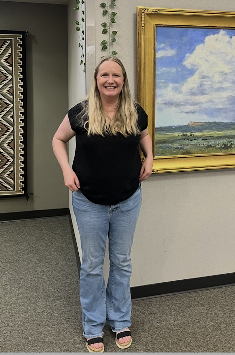 Jenny stands in the library, in front of a painting. She has long blonde hair and is wearing a black t-shirt and jeans.