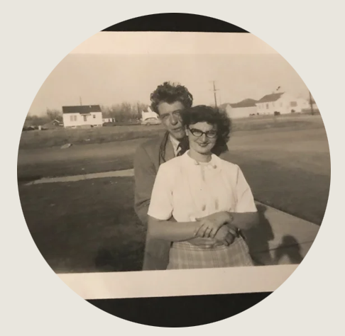 A black-and-white photo of a young couple outside. The woman smiles as the man embraces her from behind. They wear mid-century clothing. She is wearing glasses, a white blouse and skirt and he is wearing a suitcoat. Suburban houses in the background.