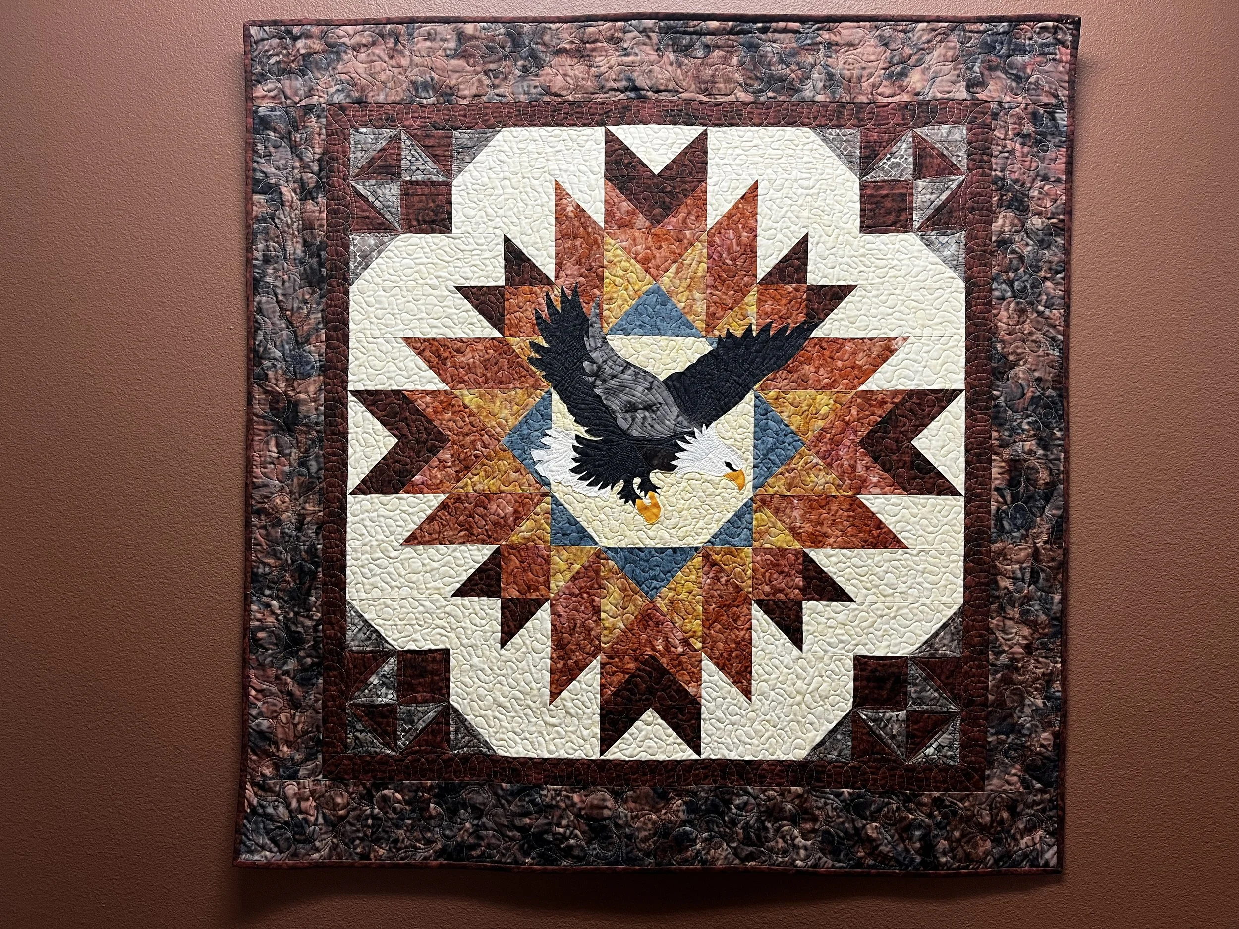 A square quilt featuring a central bald eagle in flight, surrounded by a geometric star pattern in shades of brown, cream, and blue on a wall.