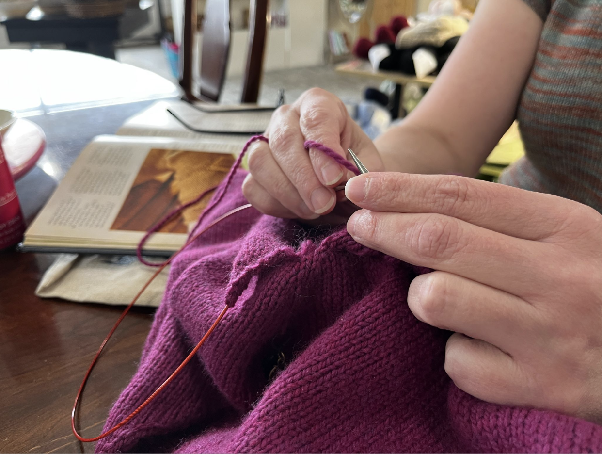 A close up photographs of hands holding a knitting needle, knitting a magenta piece of clothing. In the background there are piles of yarn and newspapers.