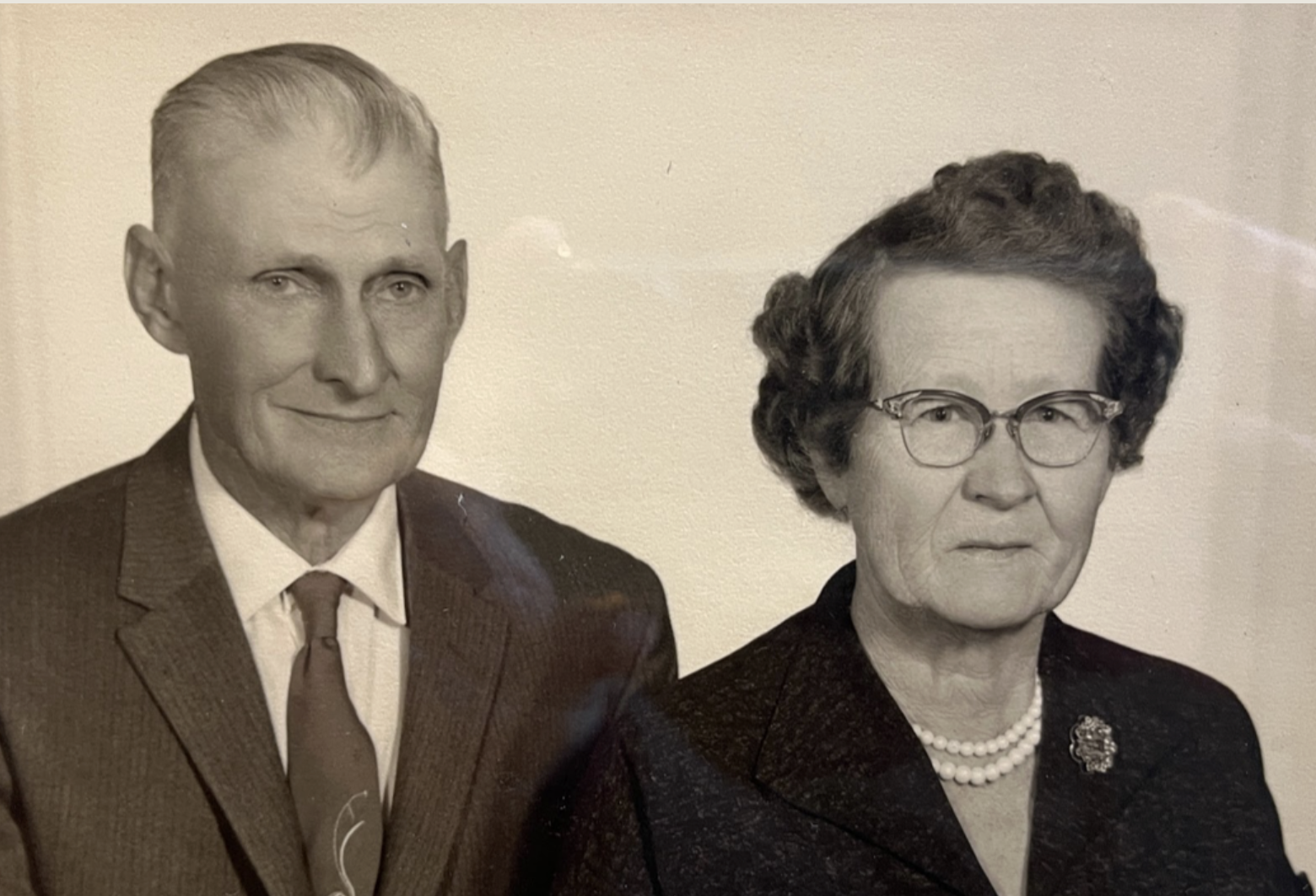 Cheyenne- Sepia-toned portrait of an older couple dressed in formal attire. The man wears a suit and tie, and the woman wears glasses and a dark jacket. Their serious expressions convey a timeless image of early 20th-century Wyoming settlers.