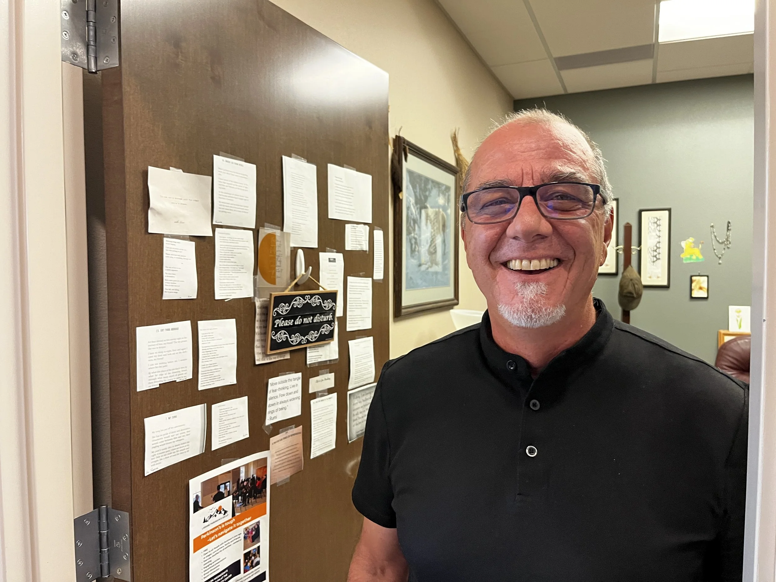 A smiling middle-aged man with glasses and a goatee stands in an office next to a wooden door covered in numerous notes and a Please Do Not Disturb sign.