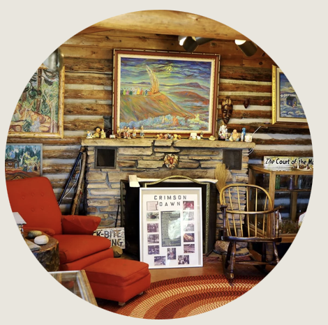 Rustic log cabin interior with stone fireplace, colorful Crimson Dawn artwork on hearth, vintage rocking chair, red armchair, and eclectic folk art decor.