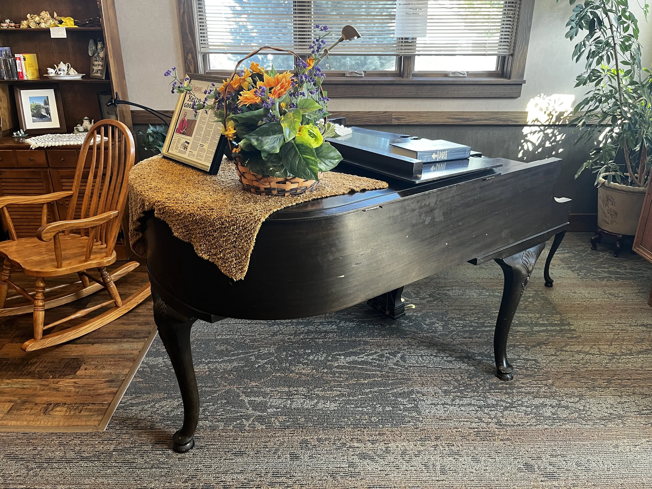Black grand piano with a flower basket, books, rocking chair, and potted plant in a sunlit room