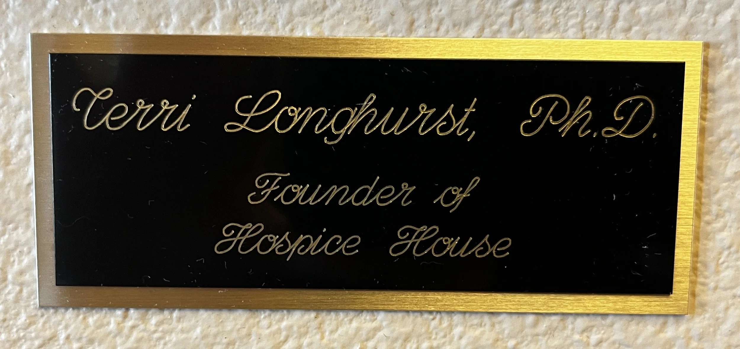 A black plaque features  gold cursive lettering that reads: Terri Longhurst, Ph.D., Founder of Hospice House, centered in two distinct lines.