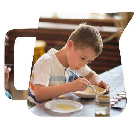 A young boy sitting at a table, painting or drawing with a brush on a plate serving as an easel, with art supplies and water jars nearby.