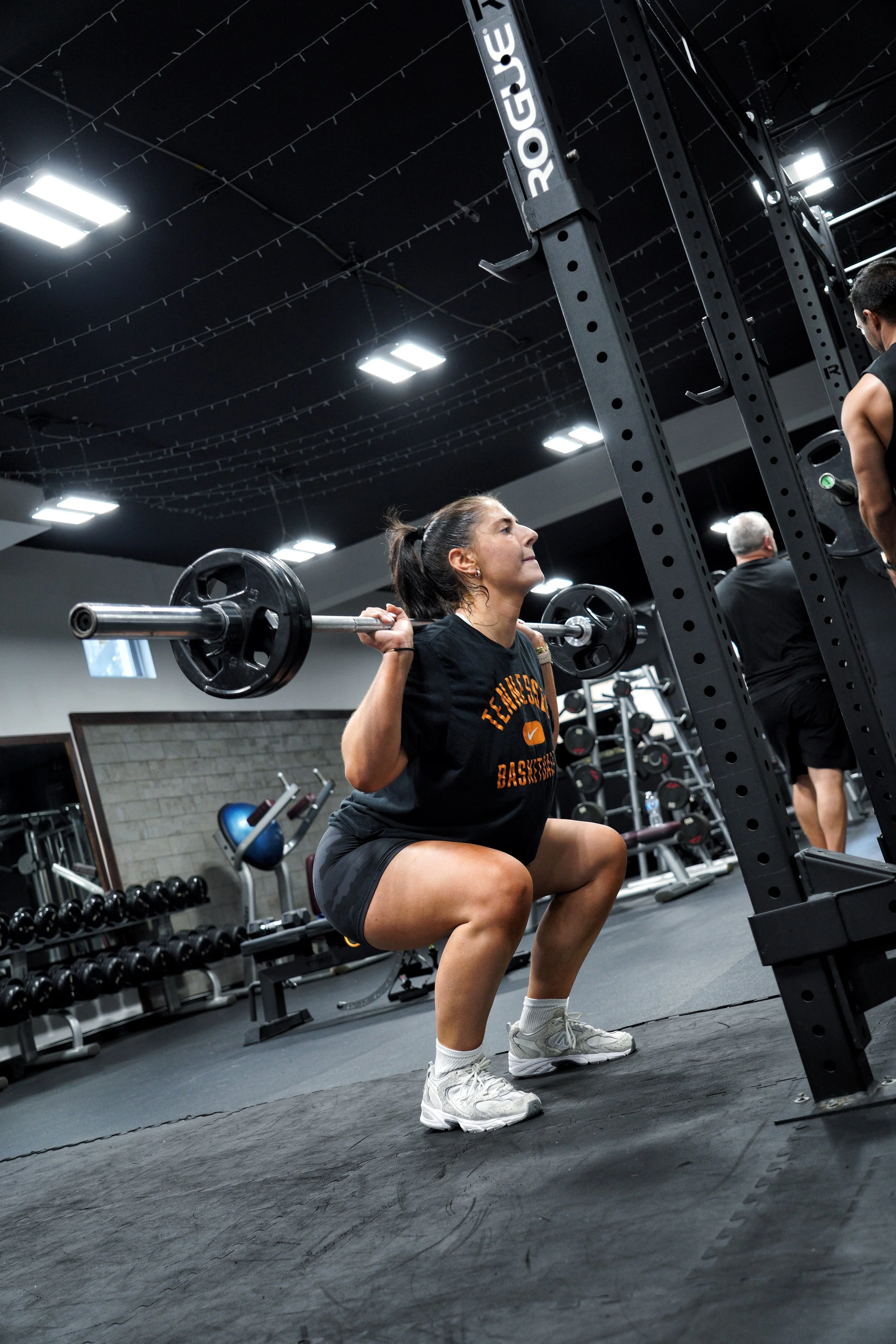 “Woman performing a barbell back squat during a training session at EMBODI Fitness, focusing on form inside the squat rack.”