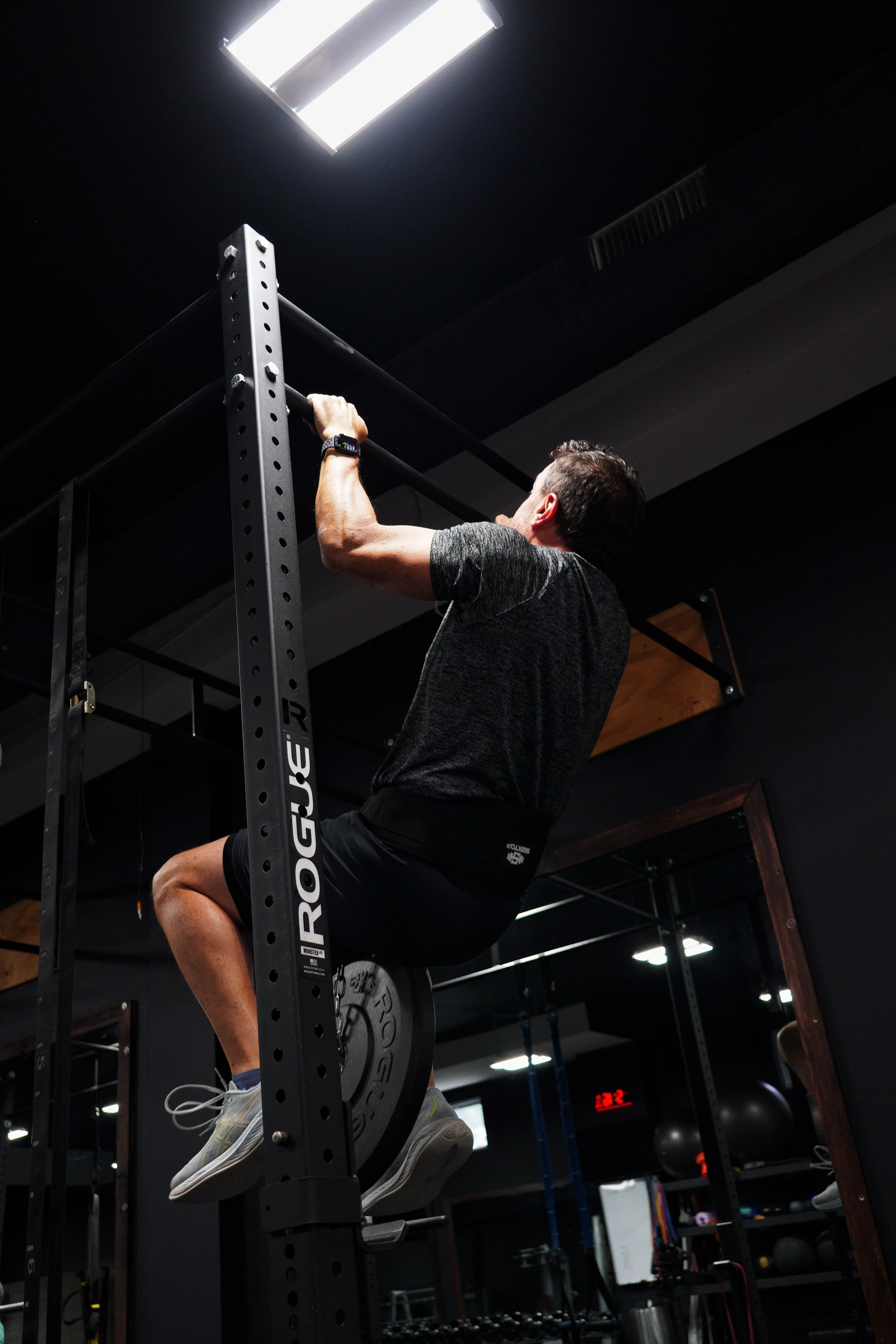 A man performing a pull-up exercise on a black power rack in a gym, with gym equipment and black walls in the background at EMBODI Fitness in Caldwell, NJ.