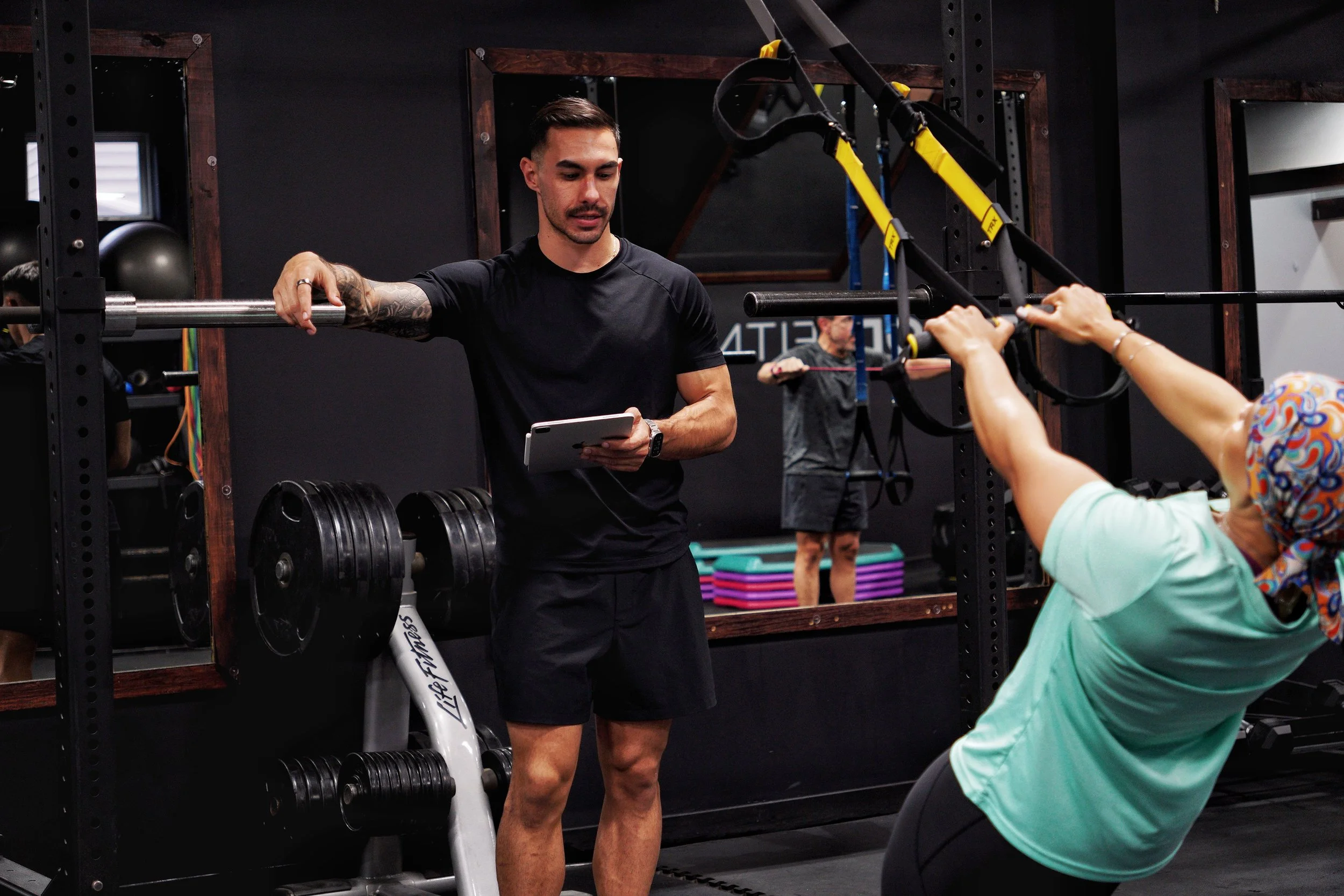 A man with tattoos in a black shirt and shorts standing by a barbell and a woman in an exercise class holding the barbell from behind. The setting is a gym with weights, mirrors, and fitness equipment.