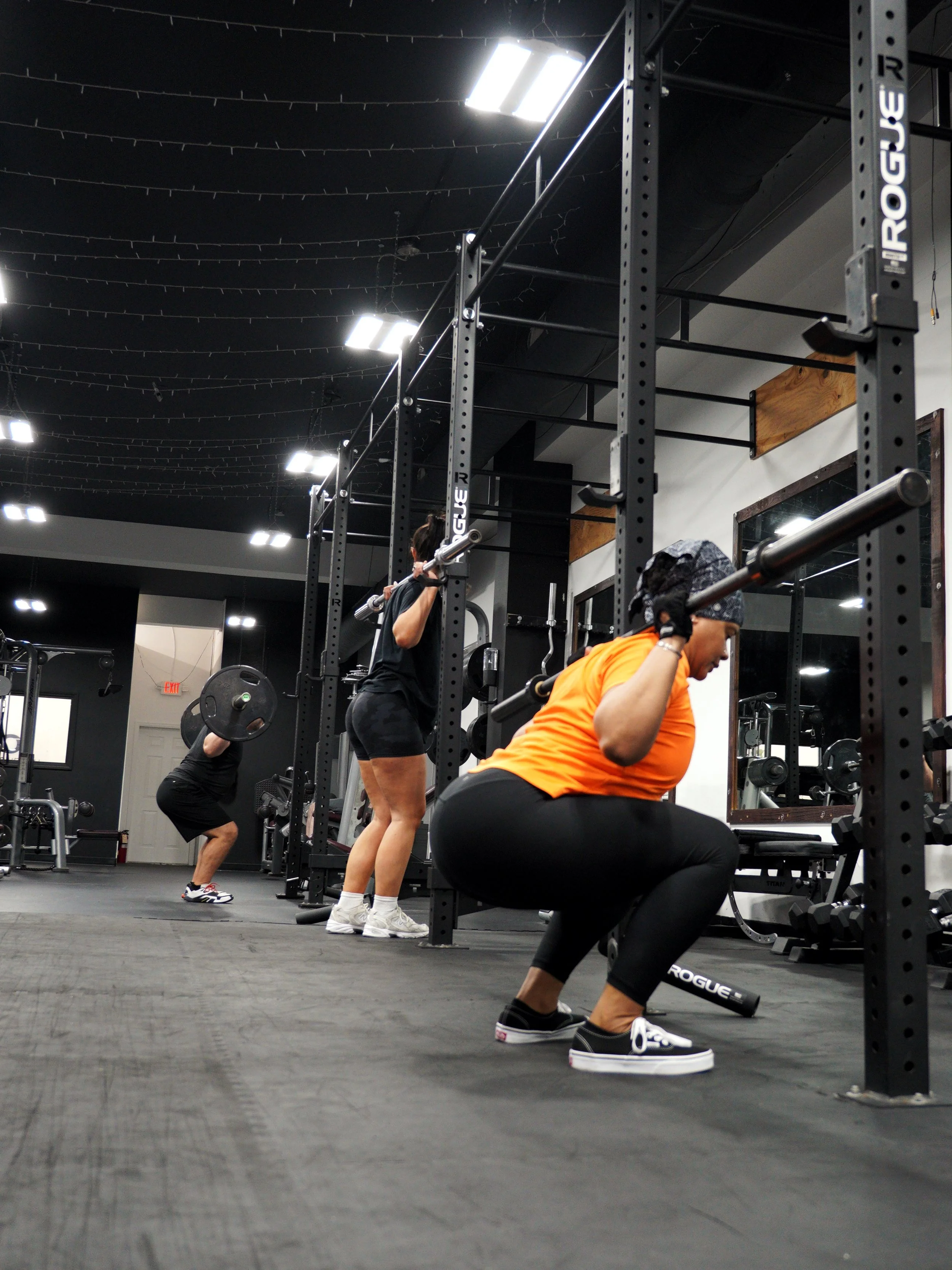 Group of clients executing barbell back squats inside the EMBODI Fitness squat racks, demonstrating structured small-group strength training.
