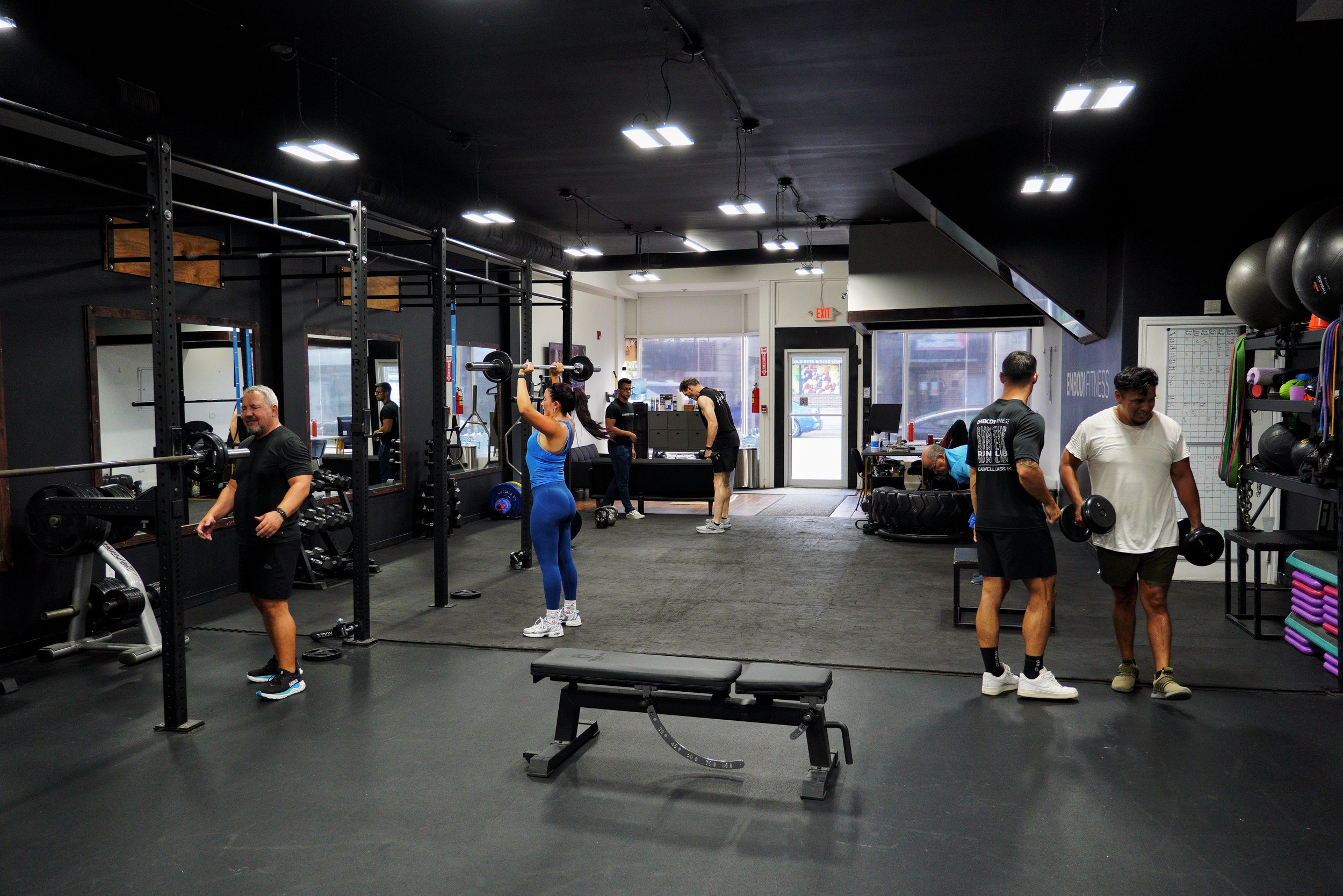 Inside a gym with several people exercising, including a woman lifting weights, men using dumbbells, and others walking or standing. Fitness equipment and workout accessories are visible, with large windows and bright ceiling lights.
