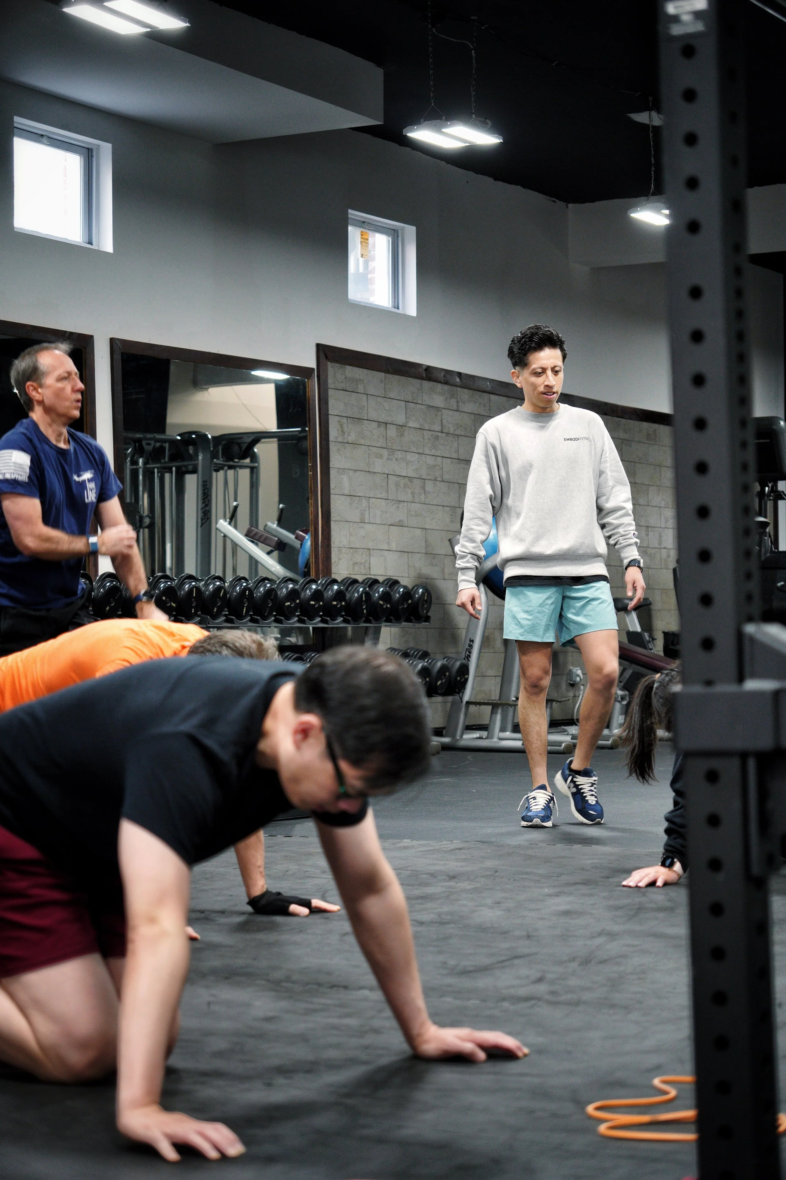 People exercising in a gym, including a man in gray sweatshirt and blue shorts walking, and others doing push-ups on the floor.
