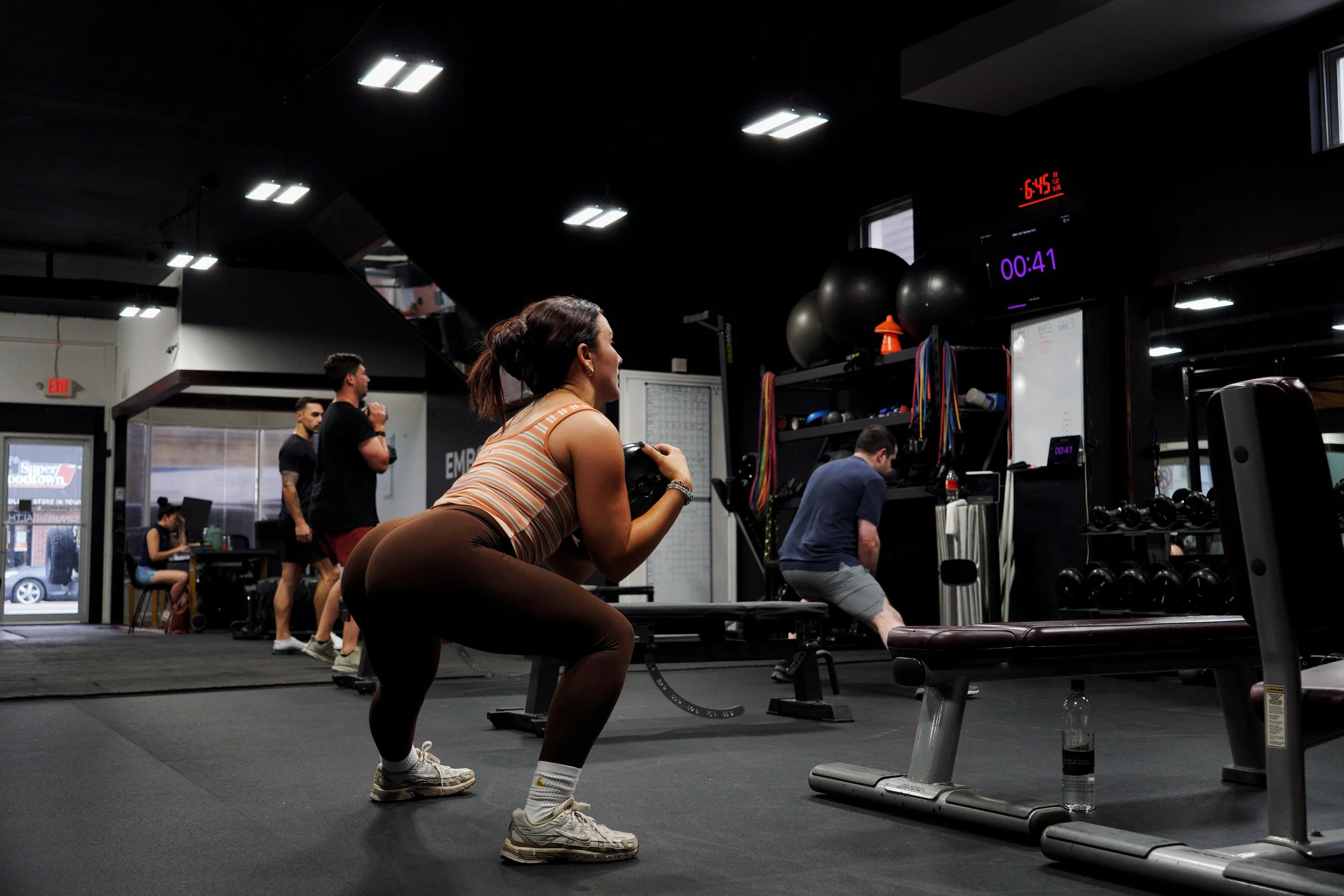 A woman with dark hair in a ponytail wearing a striped tank top and brown leggings is performing squats in a gym. There are other people working out in the background, some standing and some seated. Gym equipment, exercise balls, and a timer display are visible.