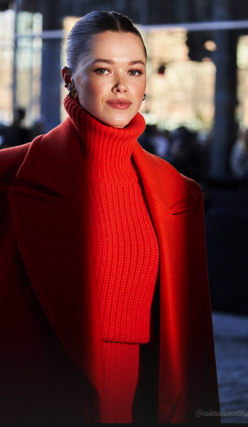 Person wearing a red turtleneck and coat, with dark hair pulled back, standing indoors.