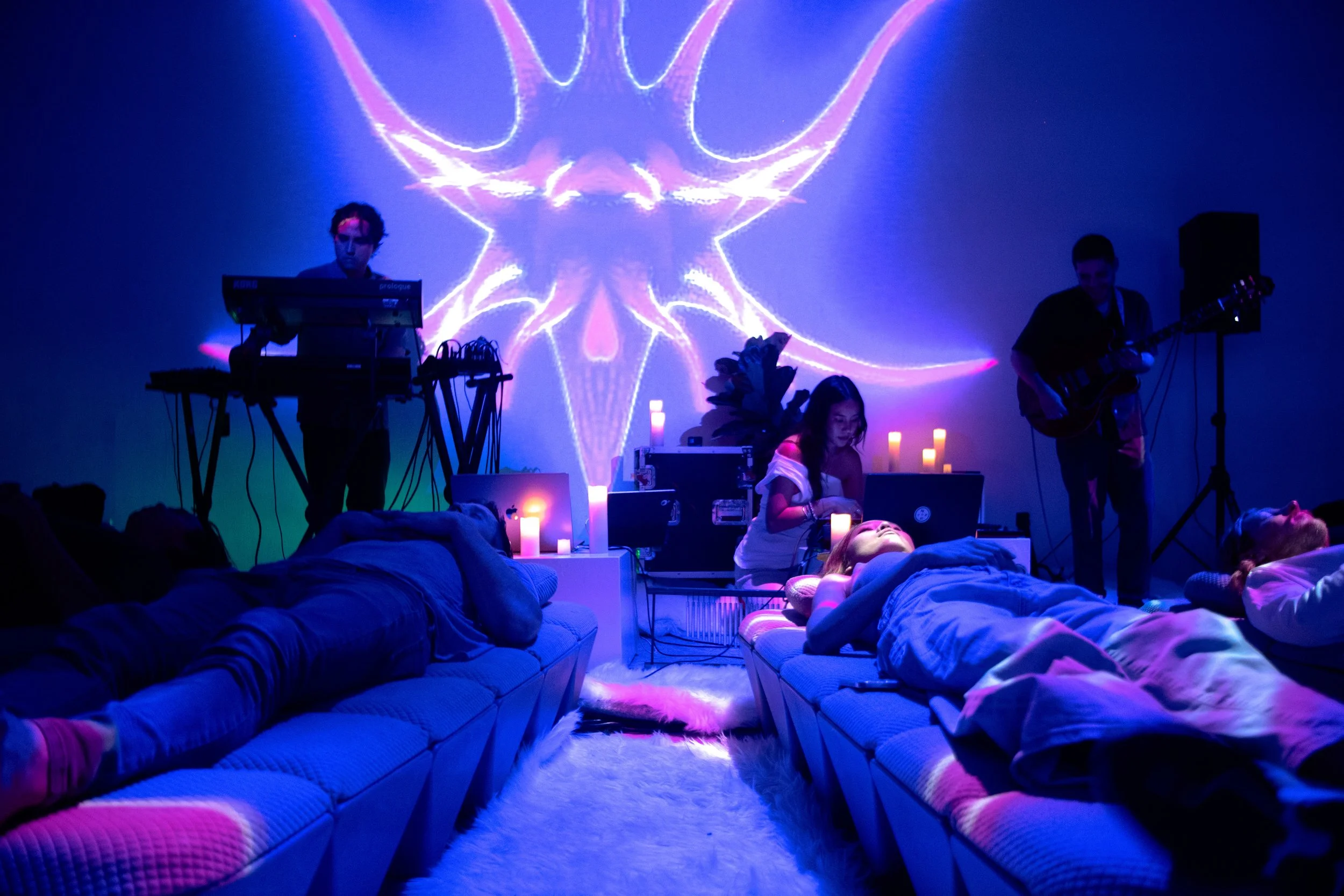 People lying on couches in a meditation or relaxation event with ambient lighting, candles, a musician, and a projection on the wall.
