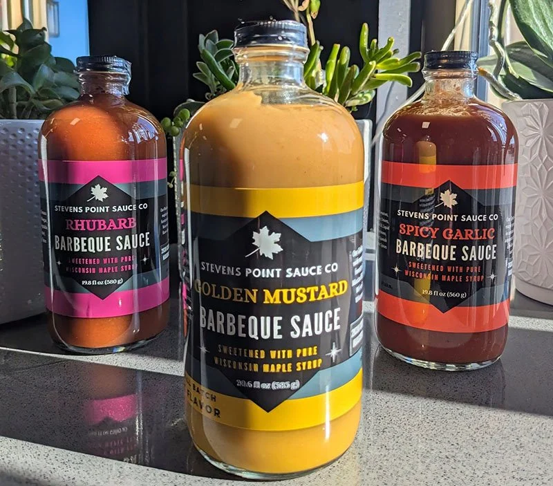 Three bottles of barbecue sauce from Stevens Point Sauce Company, with different flavors: Rhubarb, Golden Mustard, and Spicy Garlic, placed on a kitchen countertop near potted plants.