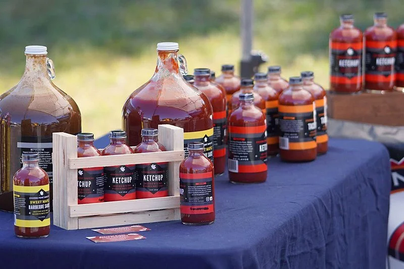 Bottles of ketchup and barbecue sauce on a table outdoors, with some in a wooden crate, at an outdoor market or event.