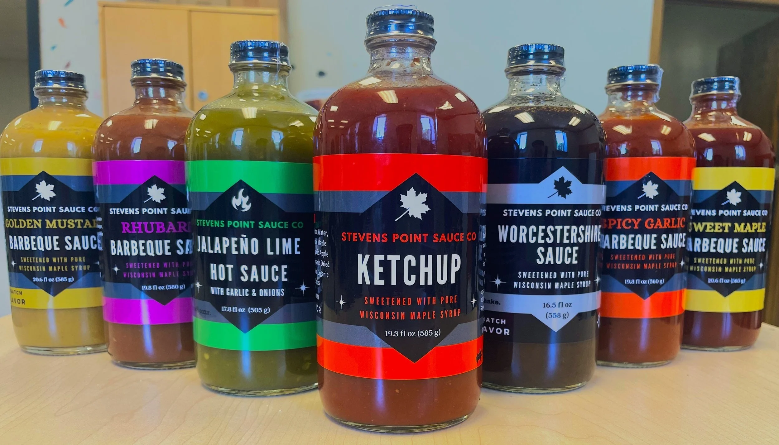 Bottles of barbecue and hot sauce in a row, featuring flavors like golden mustard, rhubarb, jalapeño lime, ketchup, Worcestershire, spicy garlic, and sweet maple, produced by Stevens Point Sauce Company