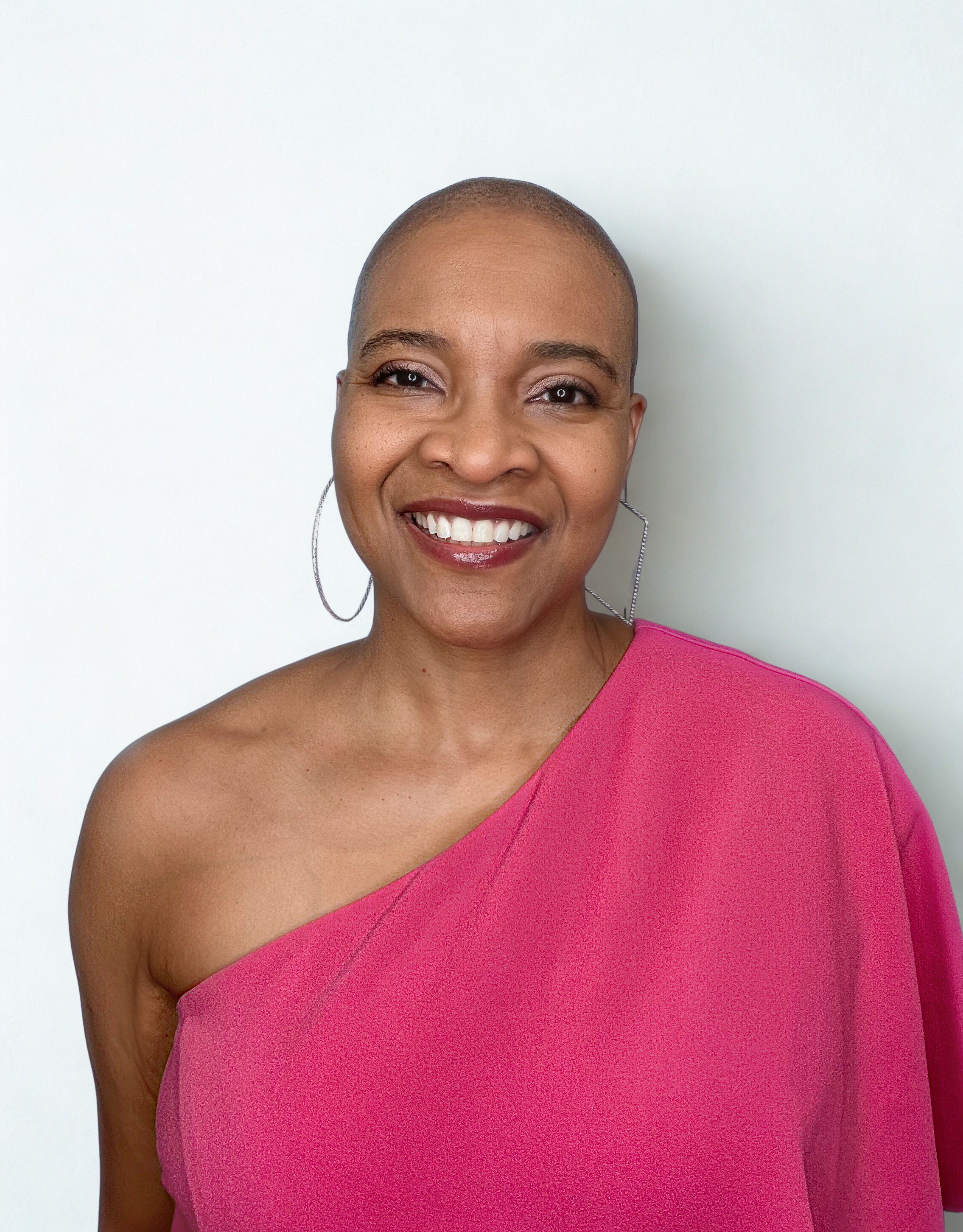 A woman with a shaved head smiling, wearing a pink one-shoulder top and hoop earrings, standing against a plain white background.