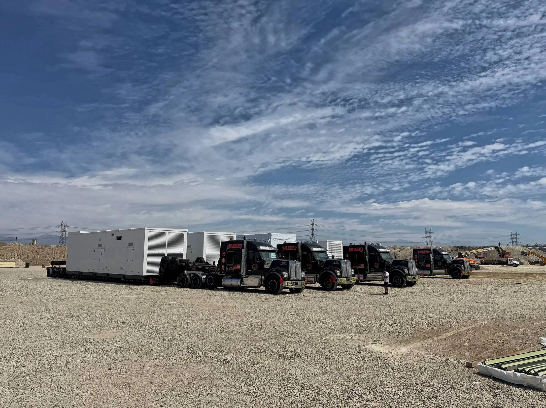 Four black heavy haul trucks lined up on a gravel site with large white generator enclosures on their trailers, power lines behind