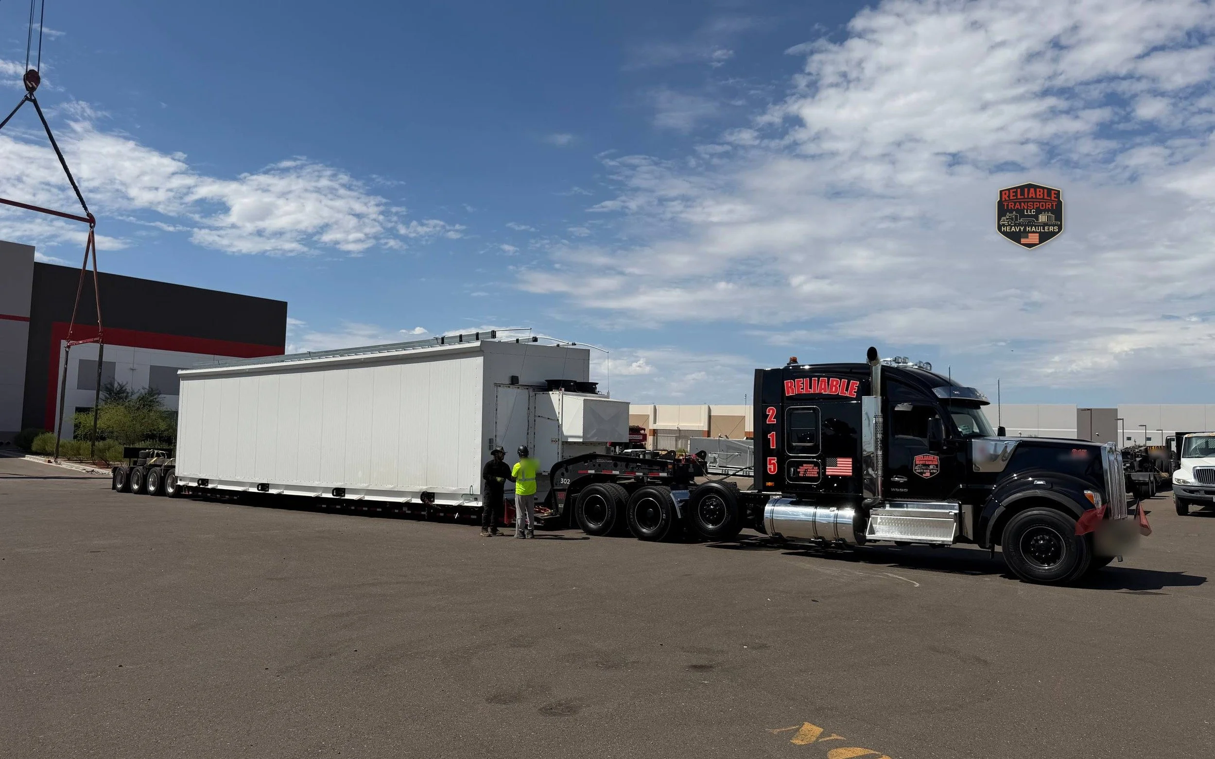 Black heavy haul tractor coupled to a white modular enclosure on a multi axle trailer under a cloudy sky