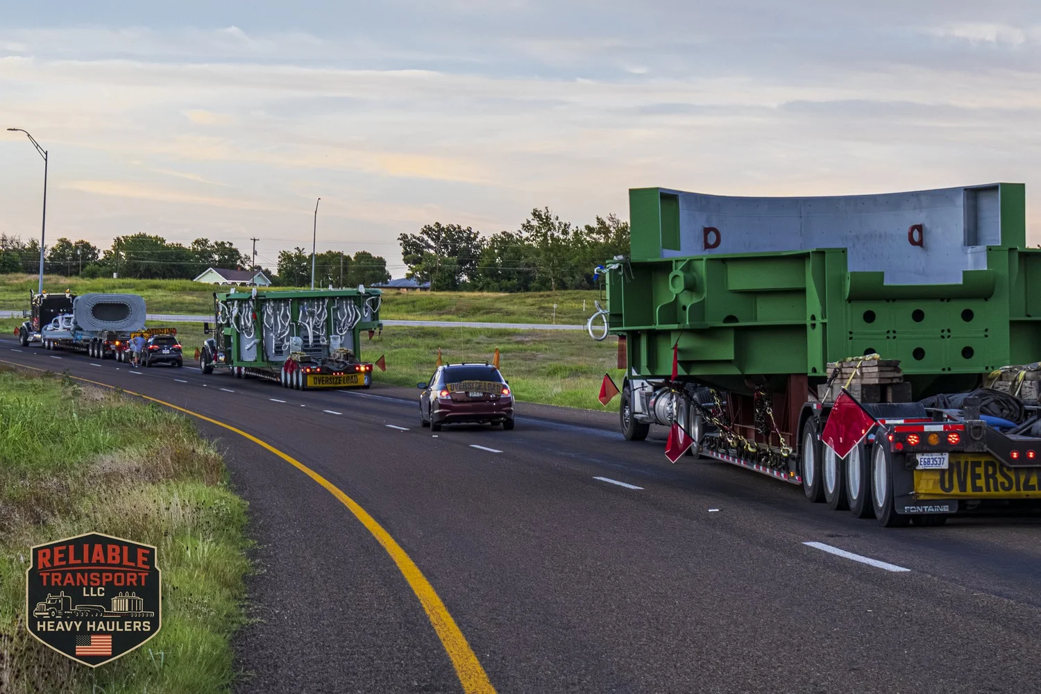 Convoy of wide loads and escort vehicles rounding a curve on a divided highway at sunset