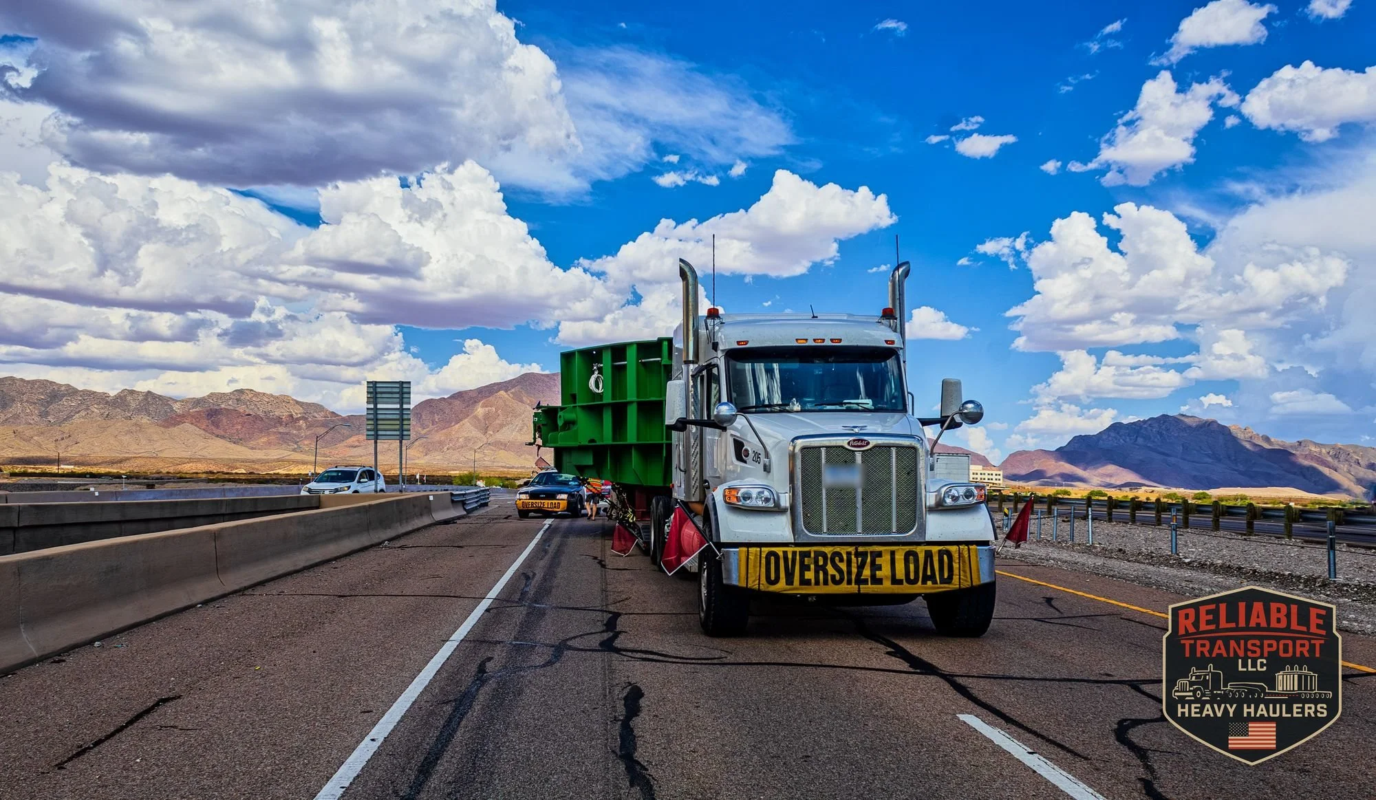 White heavy haul truck with an OVERSIZE LOAD banner pulling a wide green load on a divided desert highway, with mountains behind