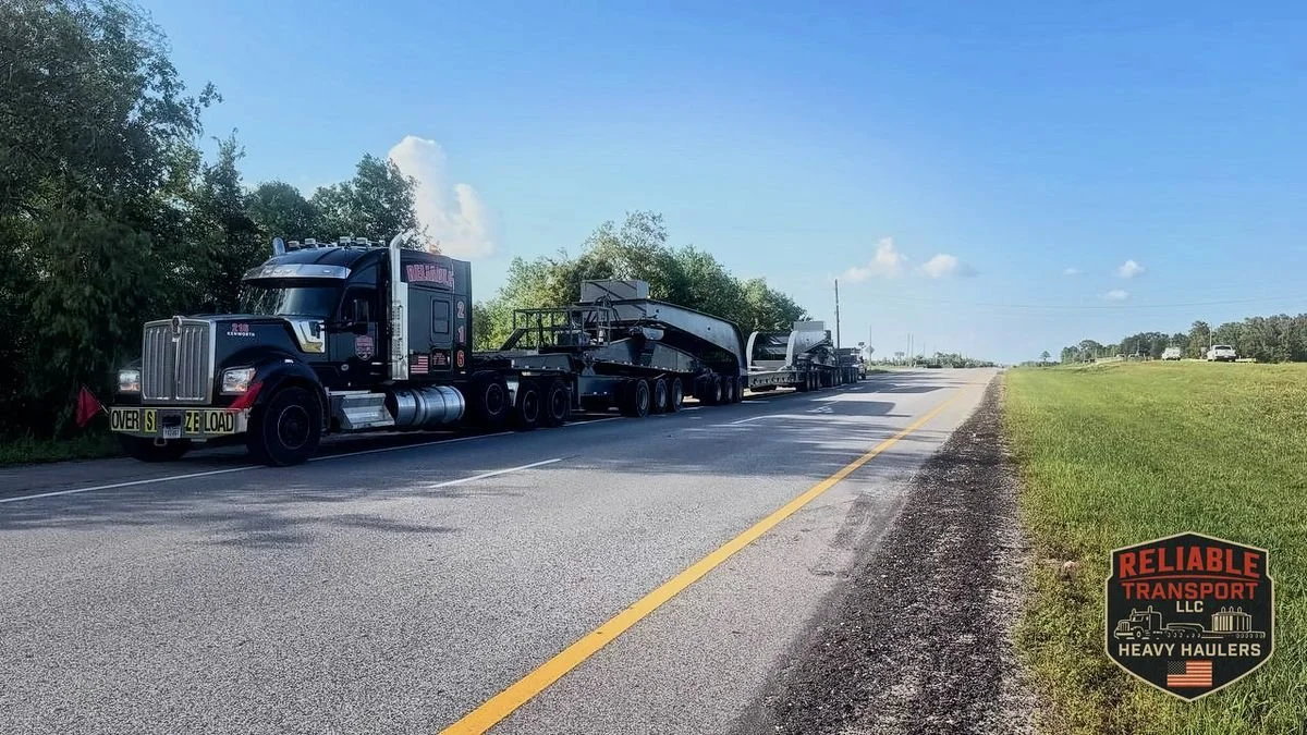 Black heavy haul truck with an OVERSIZE LOAD banner pulling an empty multi axle trailer along a two lane highway