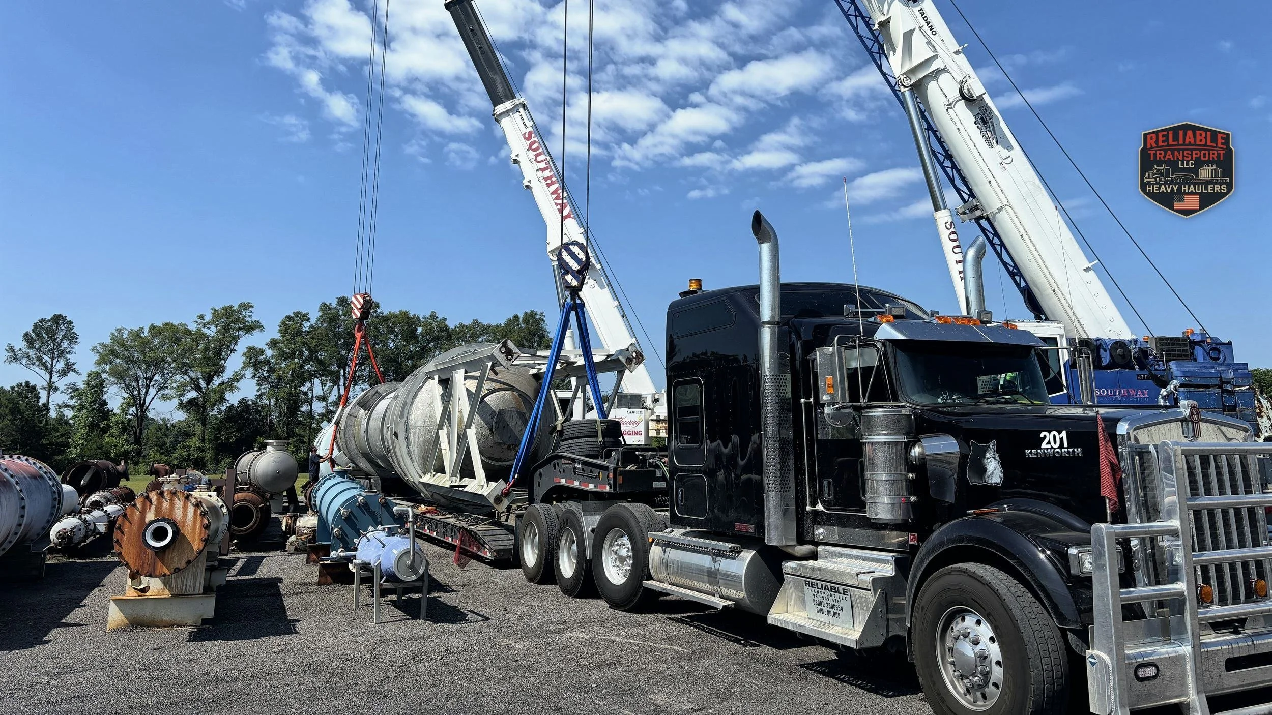 Heavy haul tractor parked by its trailer while two cranes lift a large stainless vessel