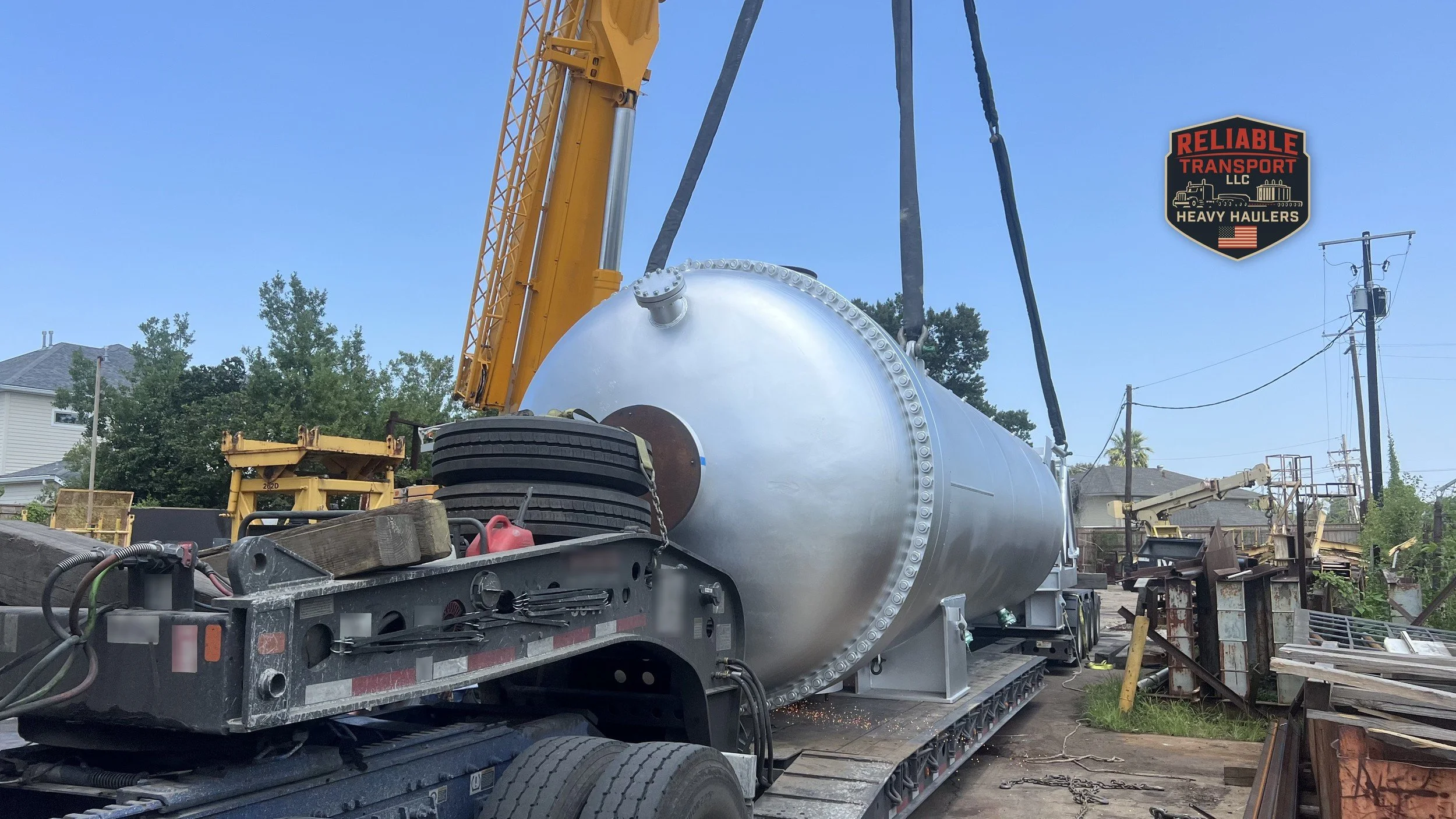 Silver pressure vessel lowered by a yellow crane onto a heavy haul trailer under a blue sky