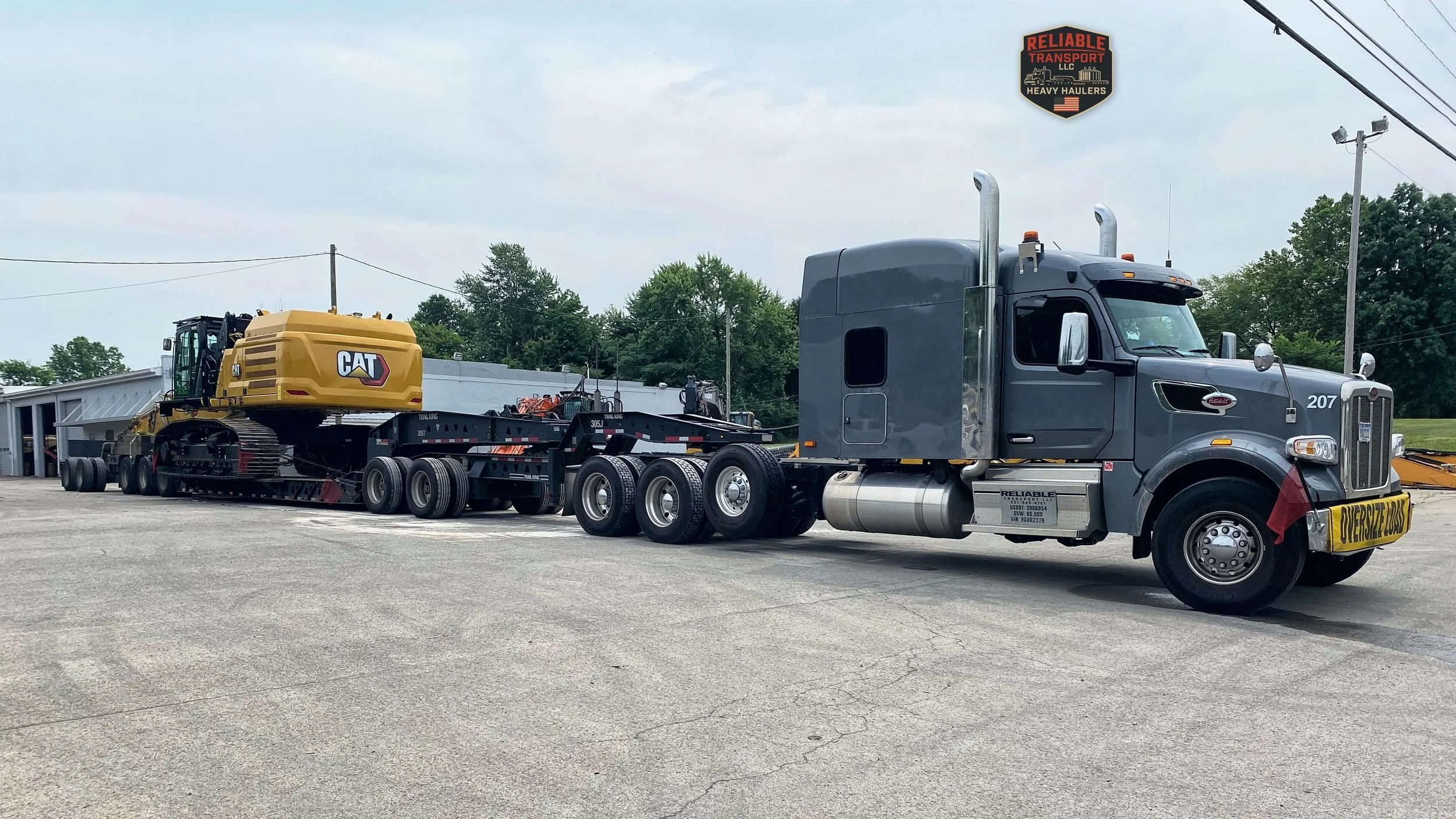 Gray heavy haul tractor with an OVERSIZE LOAD banner pulling a yellow excavator on a multi axle trailer across a paved lot