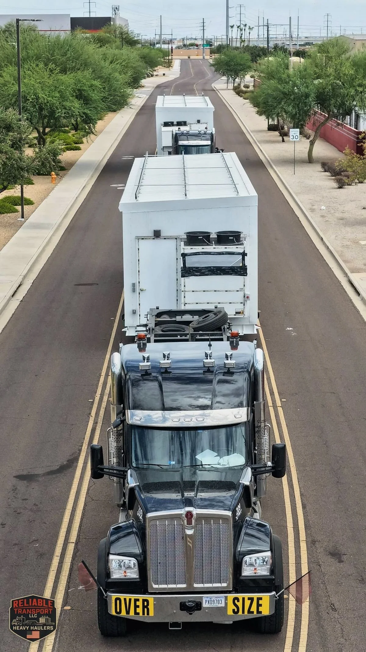 Drone view of a black truck with an OVERSIZE sign leading white module loads in convoy down a city street
