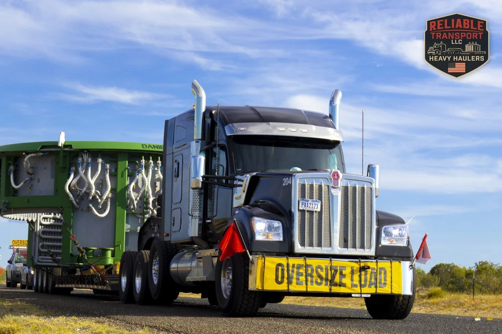 Black heavy haul tractor with red flags and an OVERSIZE LOAD banner pulling a wide green load on a sunny highway