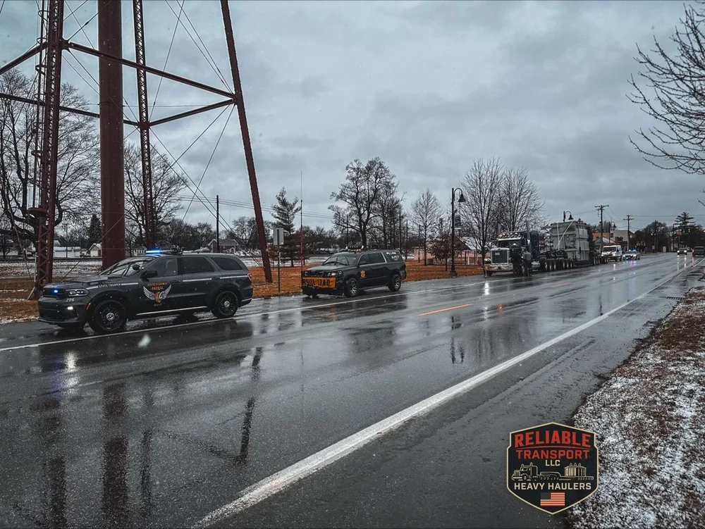 Highway patrol cruiser with its lights on and an oversize load escort vehicle ahead of the transformer load on a wet road, with snow on the roadside