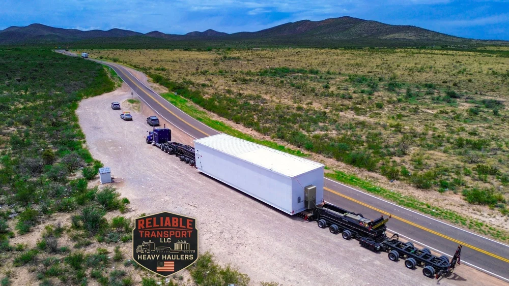 Drone view of the white switchgear building behind a blue truck, parked on the shoulder of a desert highway with escort vehicles ahead