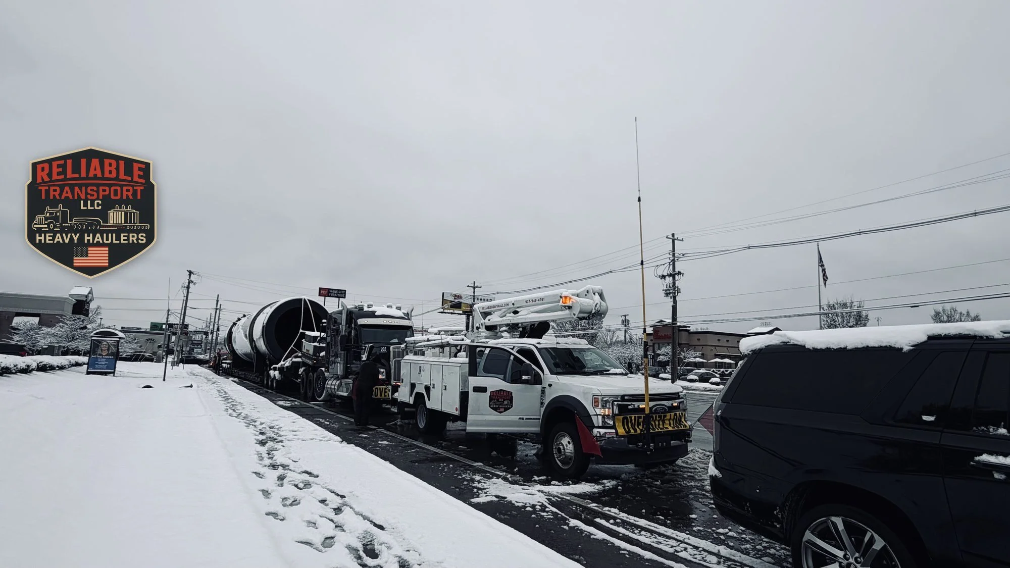 Utility bucket truck with an OVERSIZE LOAD banner stopped ahead of a heavy haul truck and a long dark drum load on a snowy street