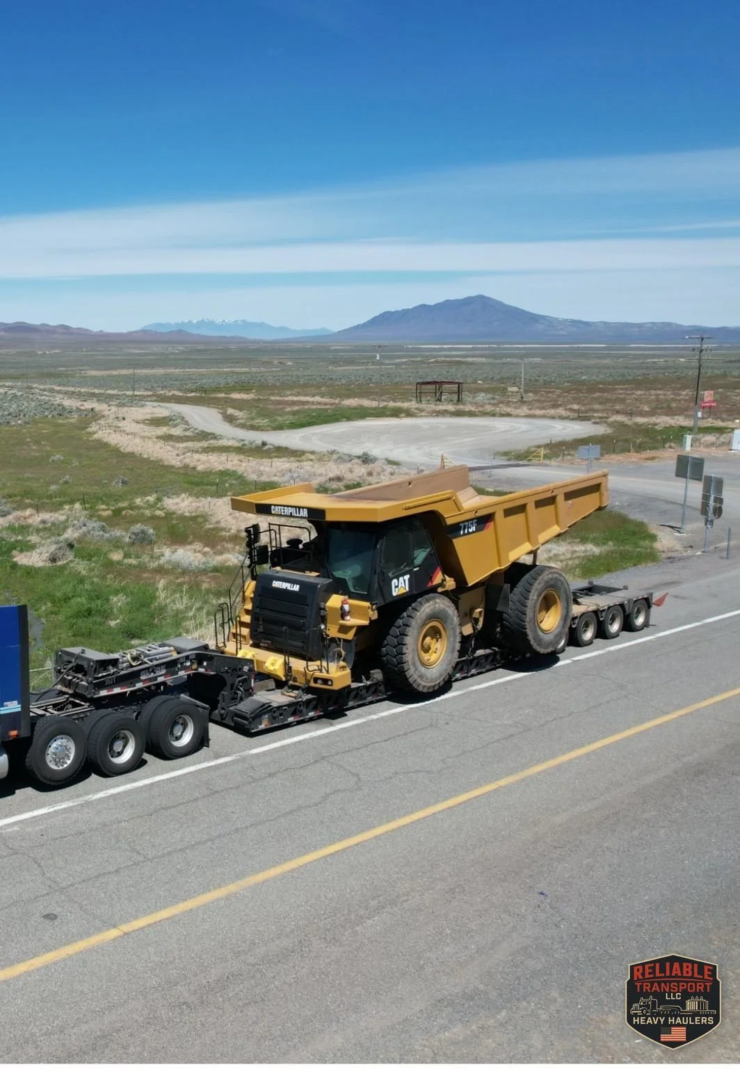 Yellow off highway haul truck on a multi axle heavy haul trailer rolling along a two lane highway, with open land and mountains behind
