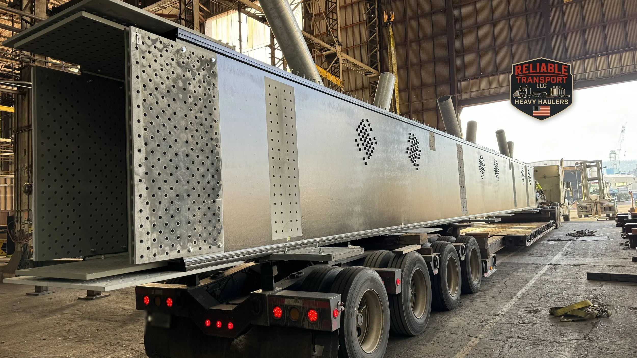 Long galvanized steel box beam on a multi axle trailer inside a large open sided industrial building