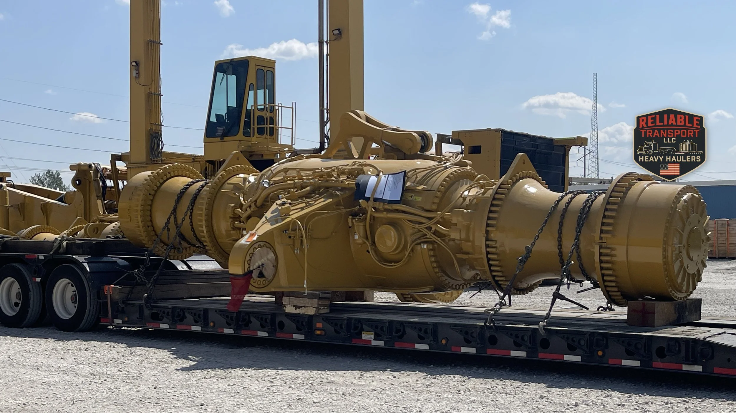 Large yellow axle assembly chained to a multi axle trailer in a gravel yard under a blue sky