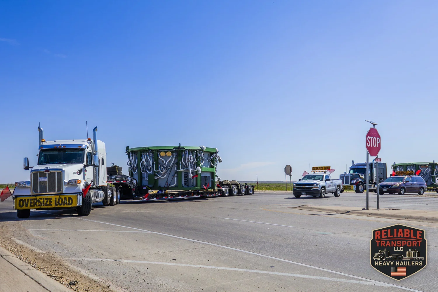 Wide load convoy crossing an intersection with a stop sign on open flat land, with an escort pickup and more wide loads following