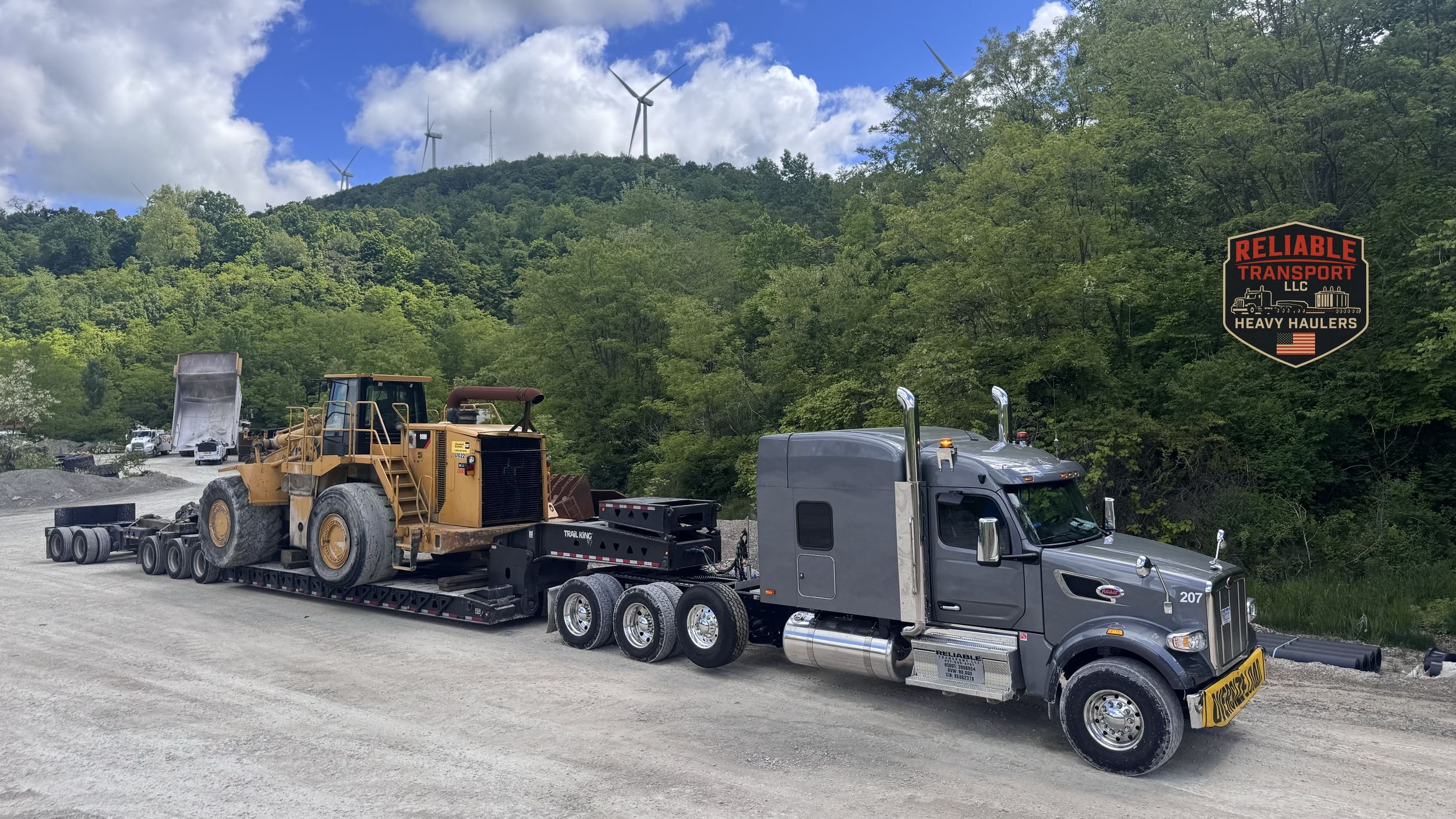 Gray heavy haul truck hauling a yellow wheel loader on a multi axle trailer in a gravel lot below green wooded hills and wind turbines