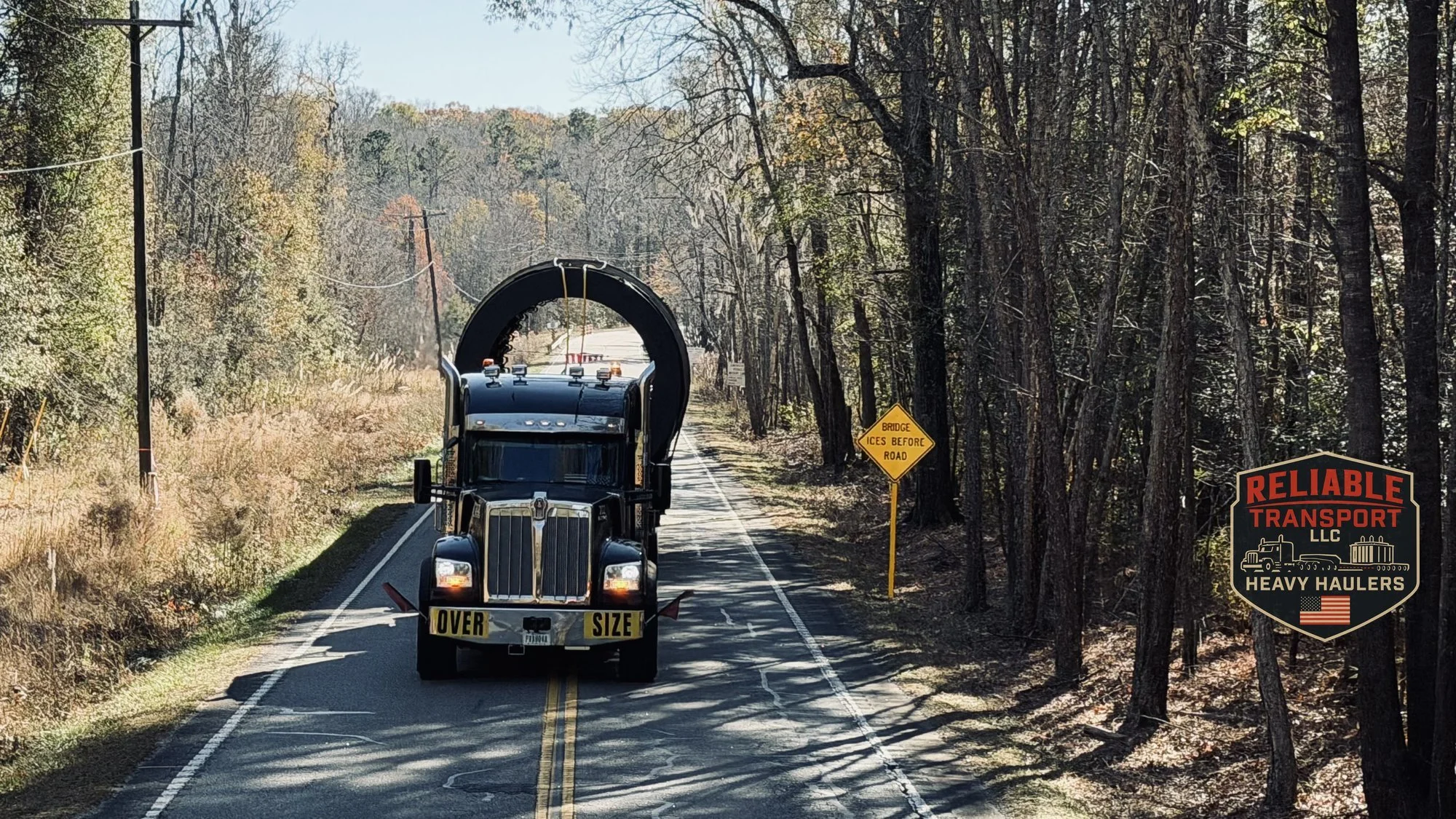 Front view of a black heavy haul truck with an OVERSIZE banner hauling a large dryer drum down a two lane road through autumn woods